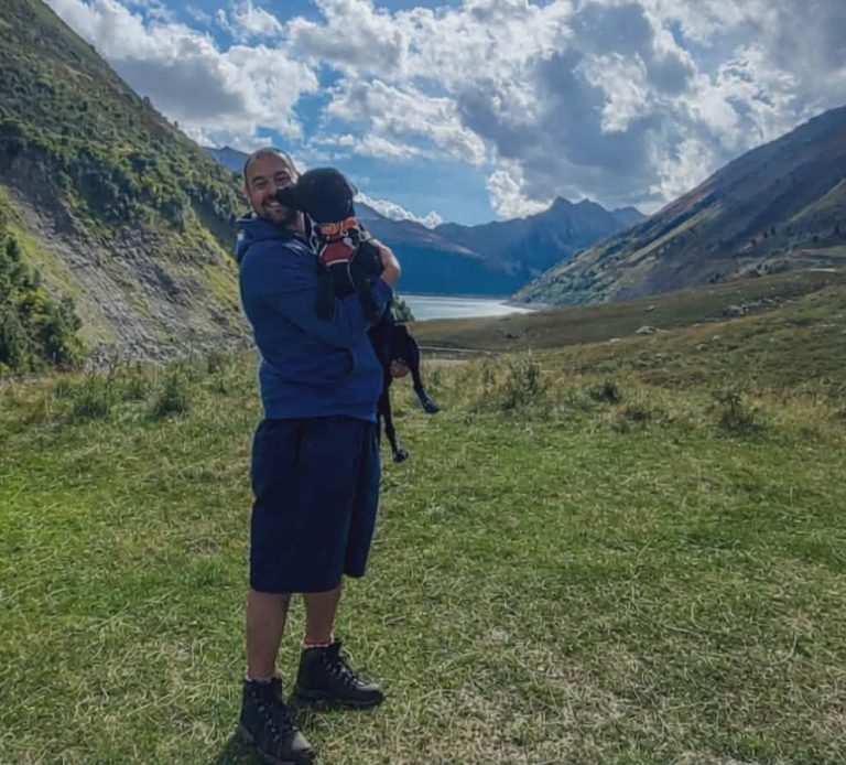 Person holding a black dog in a mountainous landscape with a lake in the background.