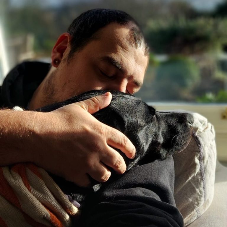 A person gently hugs a black dog while resting their forehead against it.